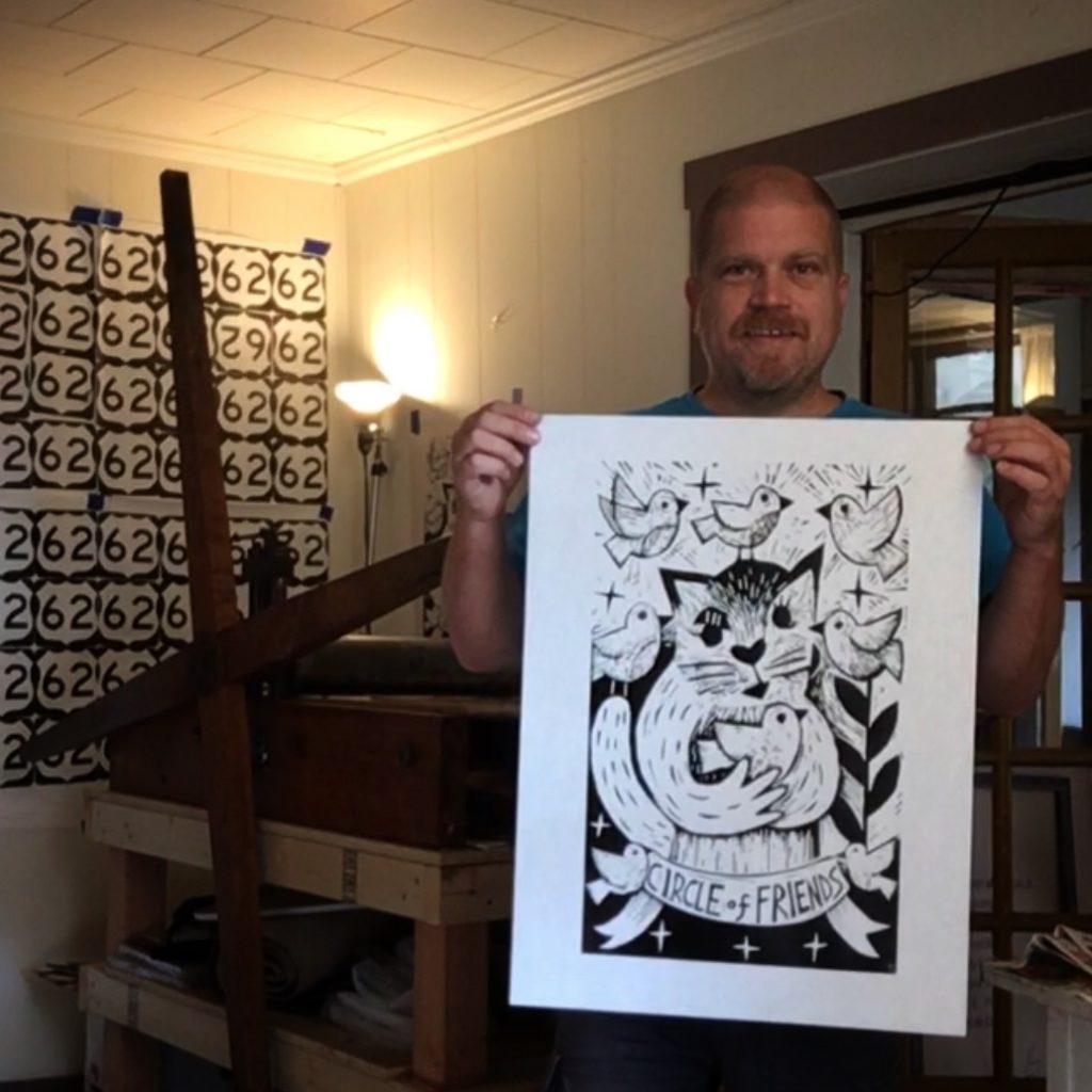 artist ken swinson holding a new woodcut of a cat holding a bird