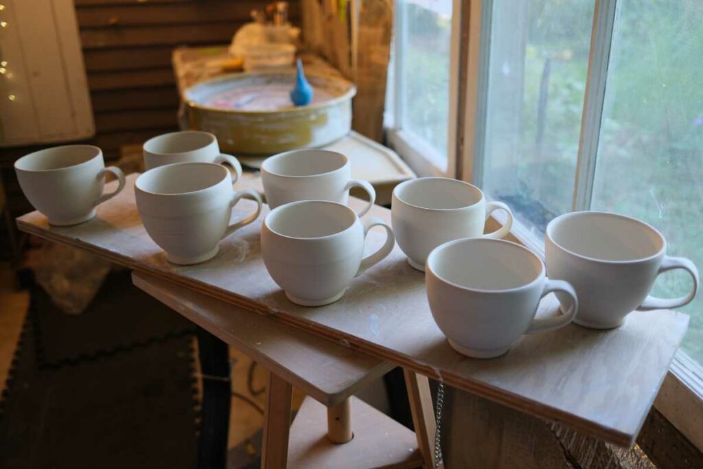 a photo of eight hand thrown porcelain cups. The handles are rather well attached, considering the artist has spent the last year or two improving his cup handles. There is a wheel in the background.