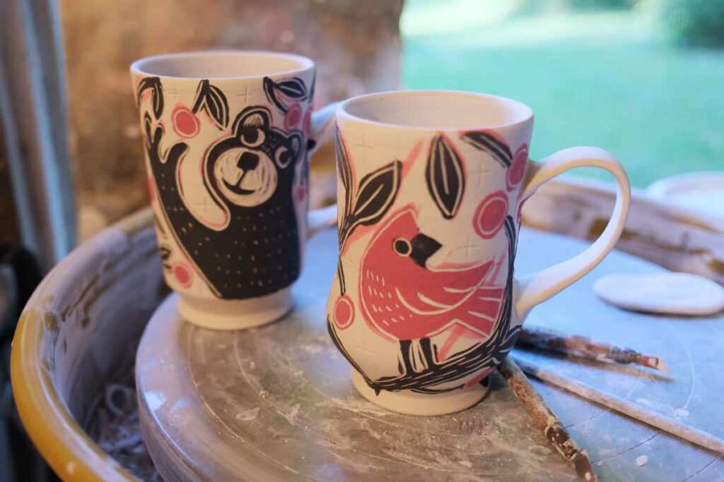 two unfired porcelain cups with a cardinal and bear picking cherries