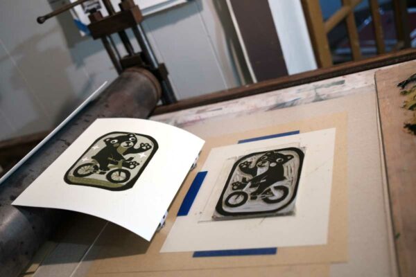 the third and final layer of a 3 layer reduction linocut by kentucky artist ken swinson. The print depicts a bear on a bike. The print is displayed beside the block. they are both on a press bed.