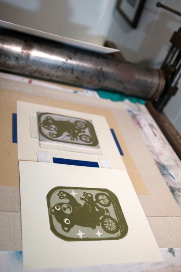 photo of work in progress linocut print displaying the second layer of a 3 color print. The silhouette of a bear on a bike is visible